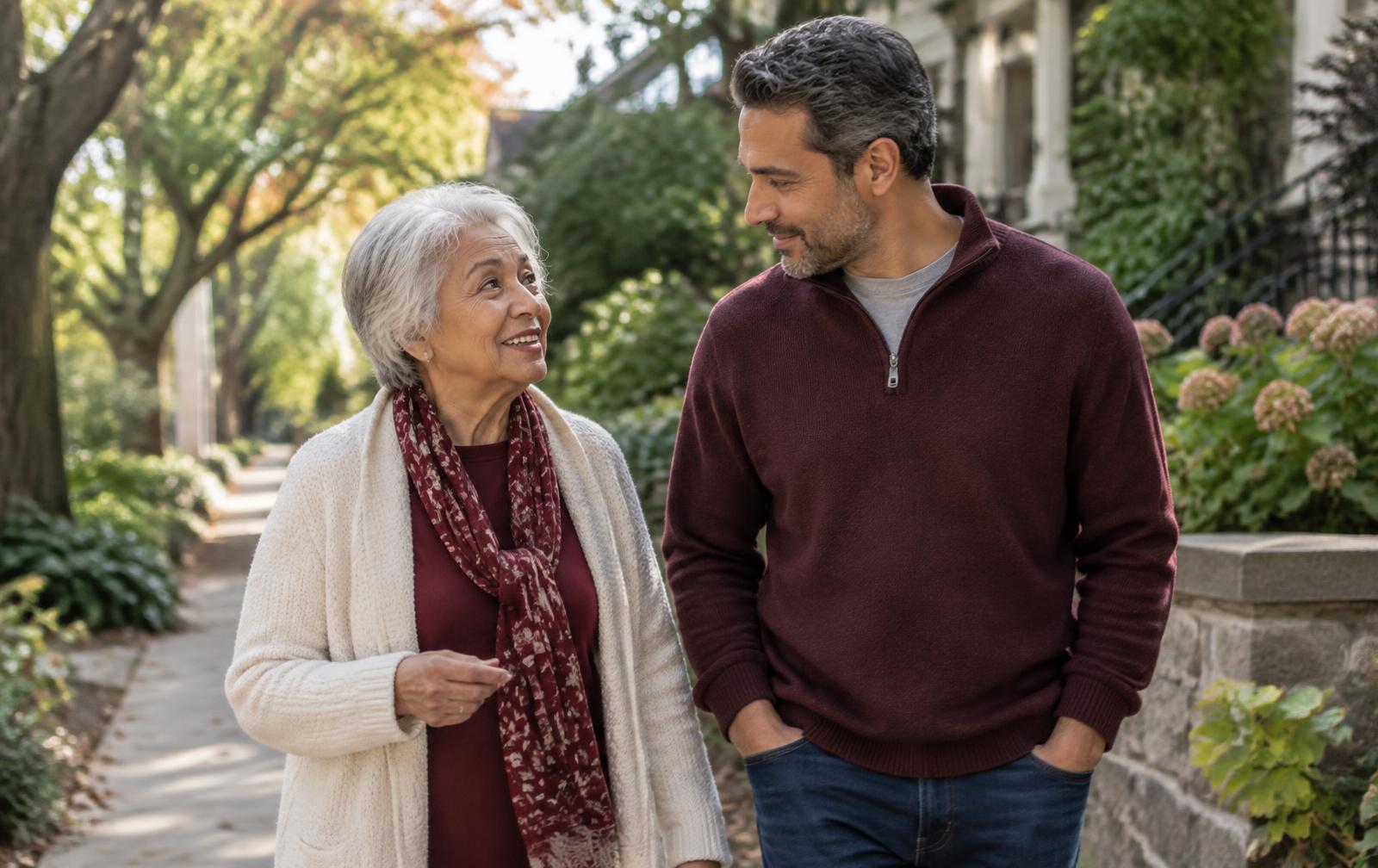 An adult son and his older mother sharing a calm conversation while walking together through a leafy neighborhood.